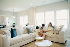 A family relaxes together on a beige sofa in a bright, modern living room with large windows and neutral decor.