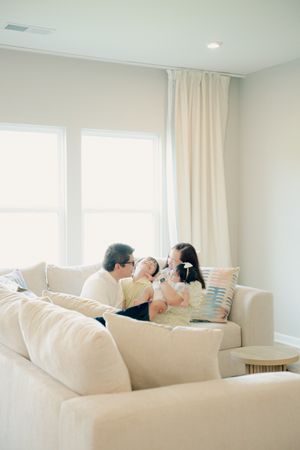 A happy family of three sits together on a cream sofa in a bright, modern living room with large windows.