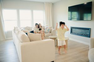 Child dancing in modern living room while family relaxes on white sofa near fireplace and wall-mounted TV.