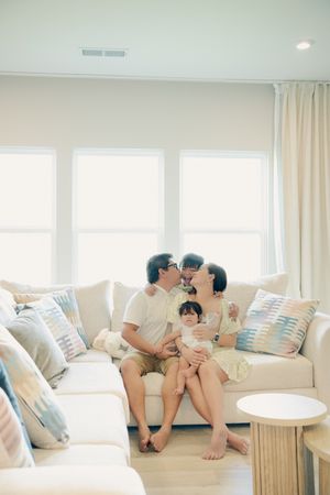 Happy Asian family of four sitting on a white sofa, parents kissing while holding their two young children.