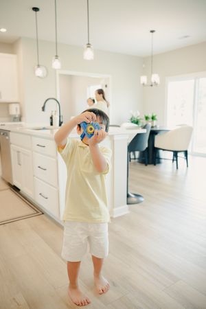 Young boy in yellow shirt photographs with blue toy camera in modern white kitchen with hardwood floors.