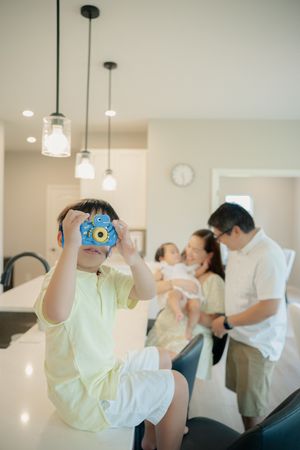 A young boy holds a blue toy camera to his face while his parents stand behind him holding a baby in a bright kitchen.