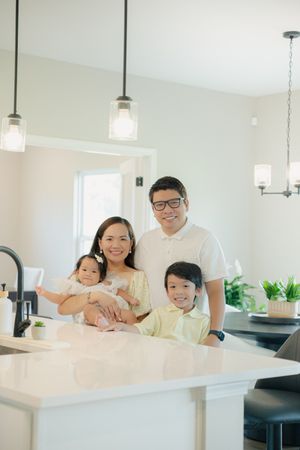 Happy Asian family of four posing in a bright modern kitchen with pendant lights and white countertops.