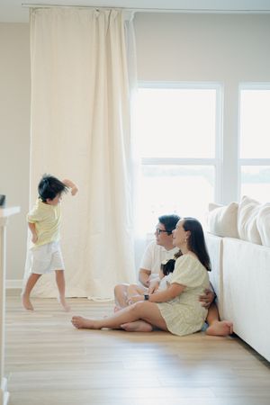 A couple sits on the floor while their young child dances near bright curtains in a modern living room.
