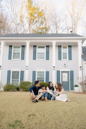Family of four sitting together on lawn in front of white colonial home with blue shutters during fall season.