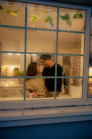 Couple sharing a romantic moment in a warmly lit kitchen, viewed through a white-framed window at dusk.