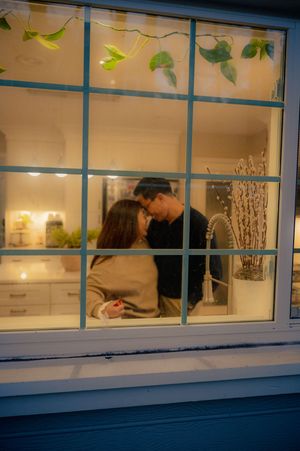 Couple sharing a romantic moment by a kitchen window, viewed from outside at night with warm interior lighting.