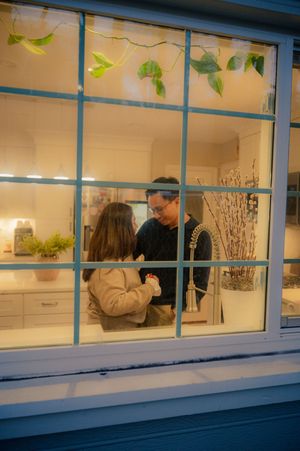Two people sharing a warm moment in a cozy kitchen, viewed through a white-framed window at night.