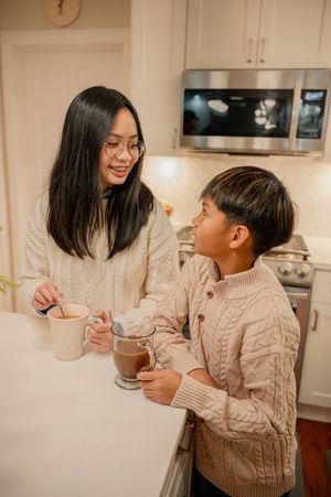 A mother and child in cozy knit sweaters share warm drinks at a kitchen counter, smiling at each other.