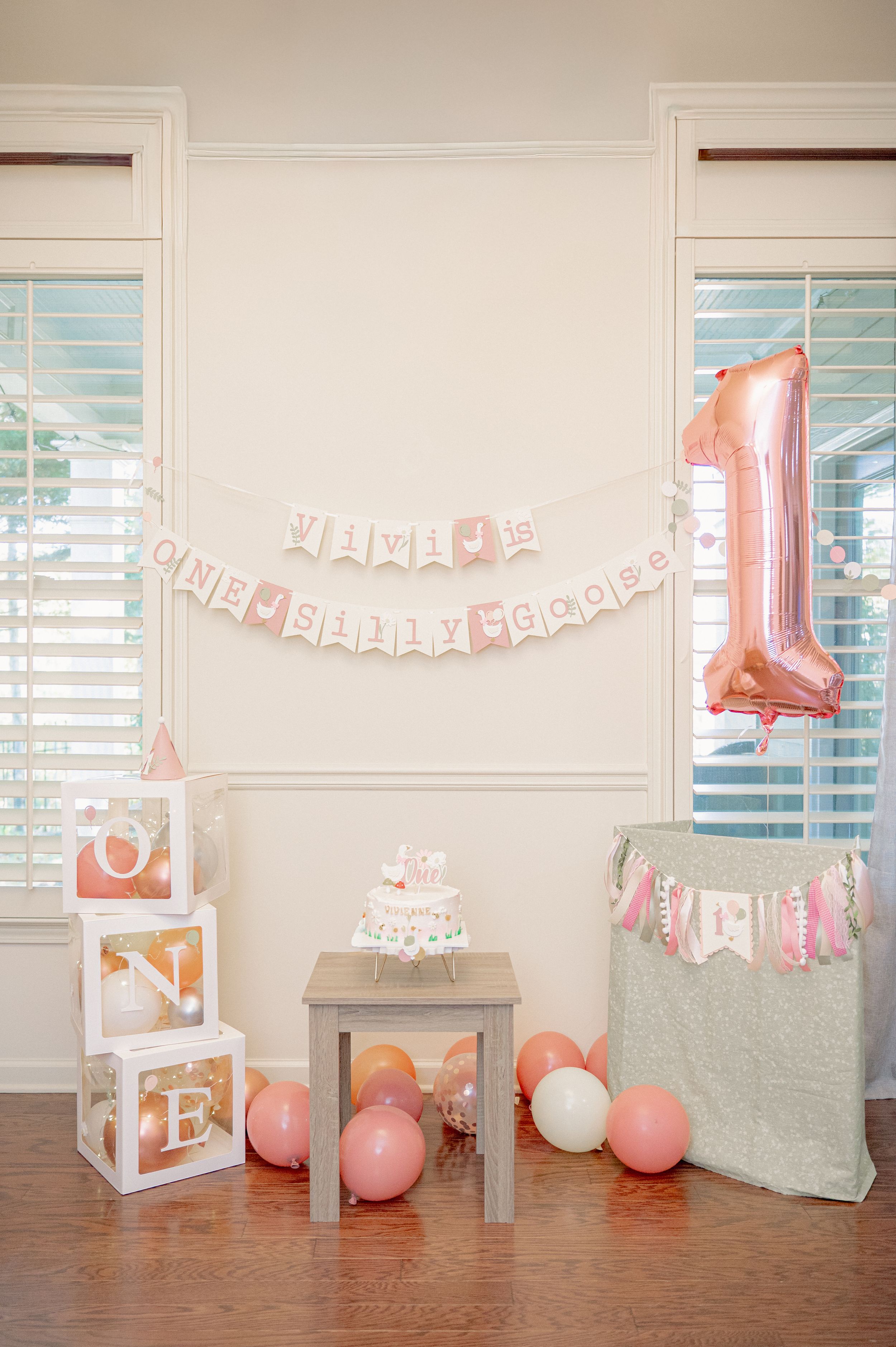 Pink and gold first birthday party setup with ONE blocks, balloon number 1, small cake, and festive banner decorations.