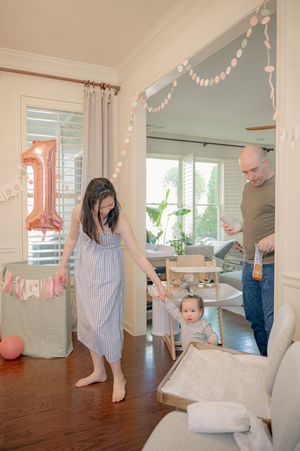 Family celebrating baby's first birthday with number one balloon, garland decorations, and parents helping toddler walk.