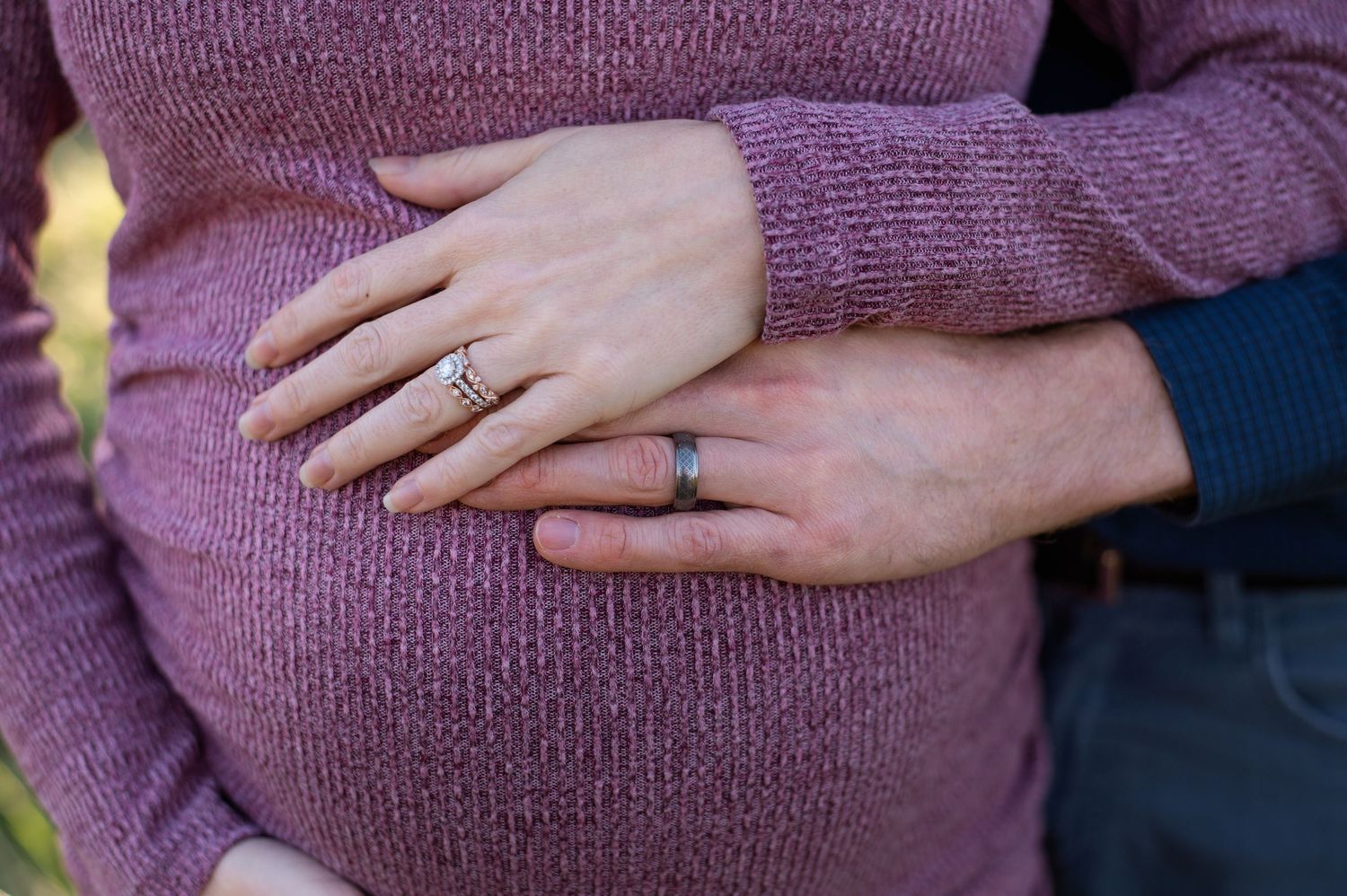 Pregnant woman in purple sweater with husband's hands on baby bump, both wearing wedding rings.