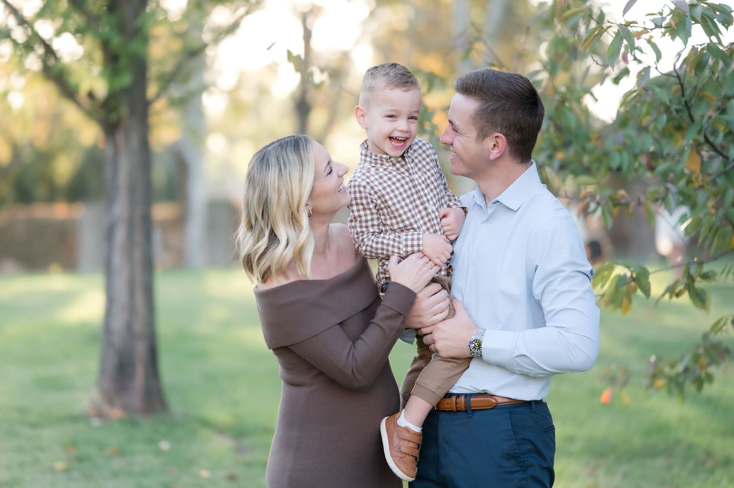 Happy family portrait with parents holding laughing toddler outdoors during golden hour.
