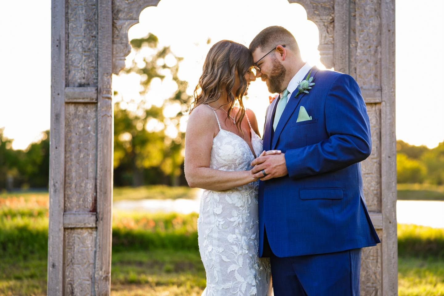 Bride and groom share a romantic moment under a rustic arch at golden hour during an outdoor wedding ceremony.