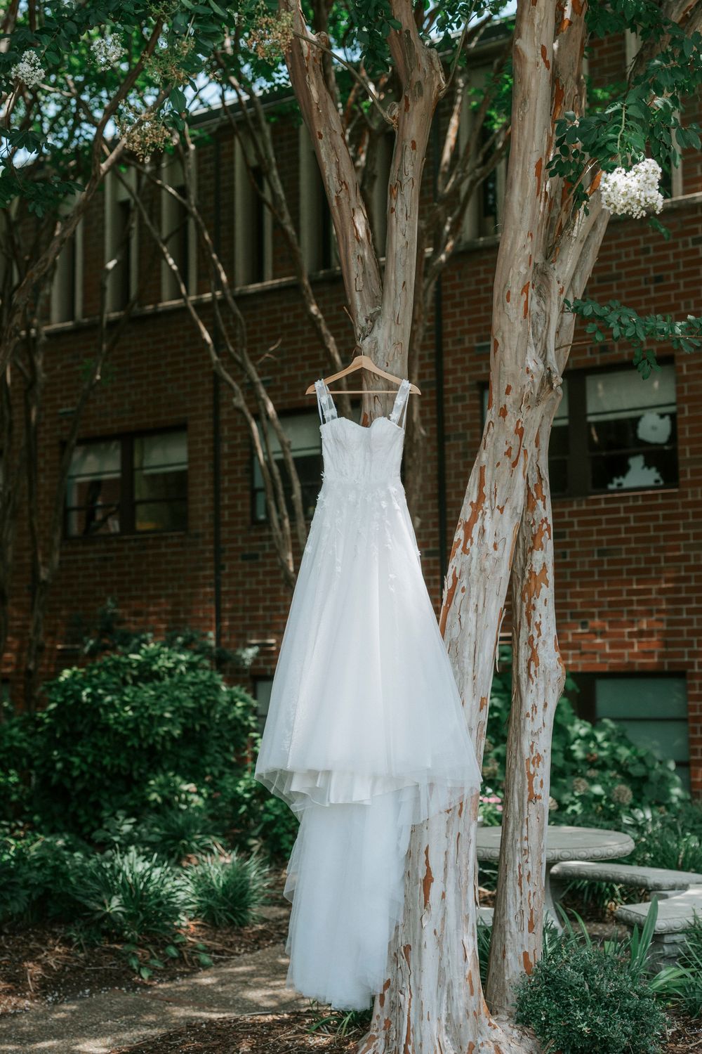 A white wedding gown with floral lace bodice hangs from a tree outside a brick building.