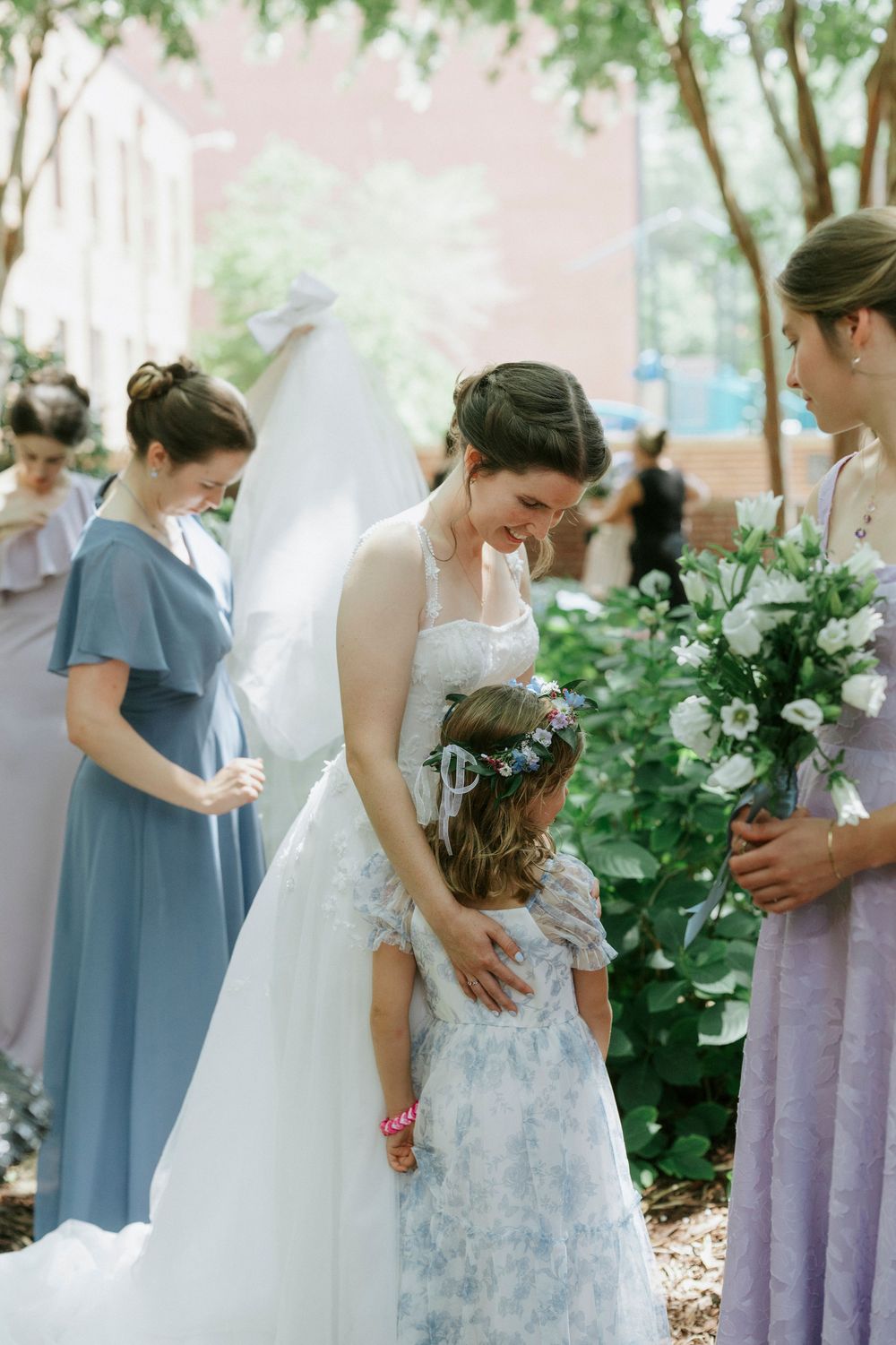 Bride in white gown smiles at flower girl in blue floral dress, surrounded by bridesmaids in soft blue and lavender outdoors.