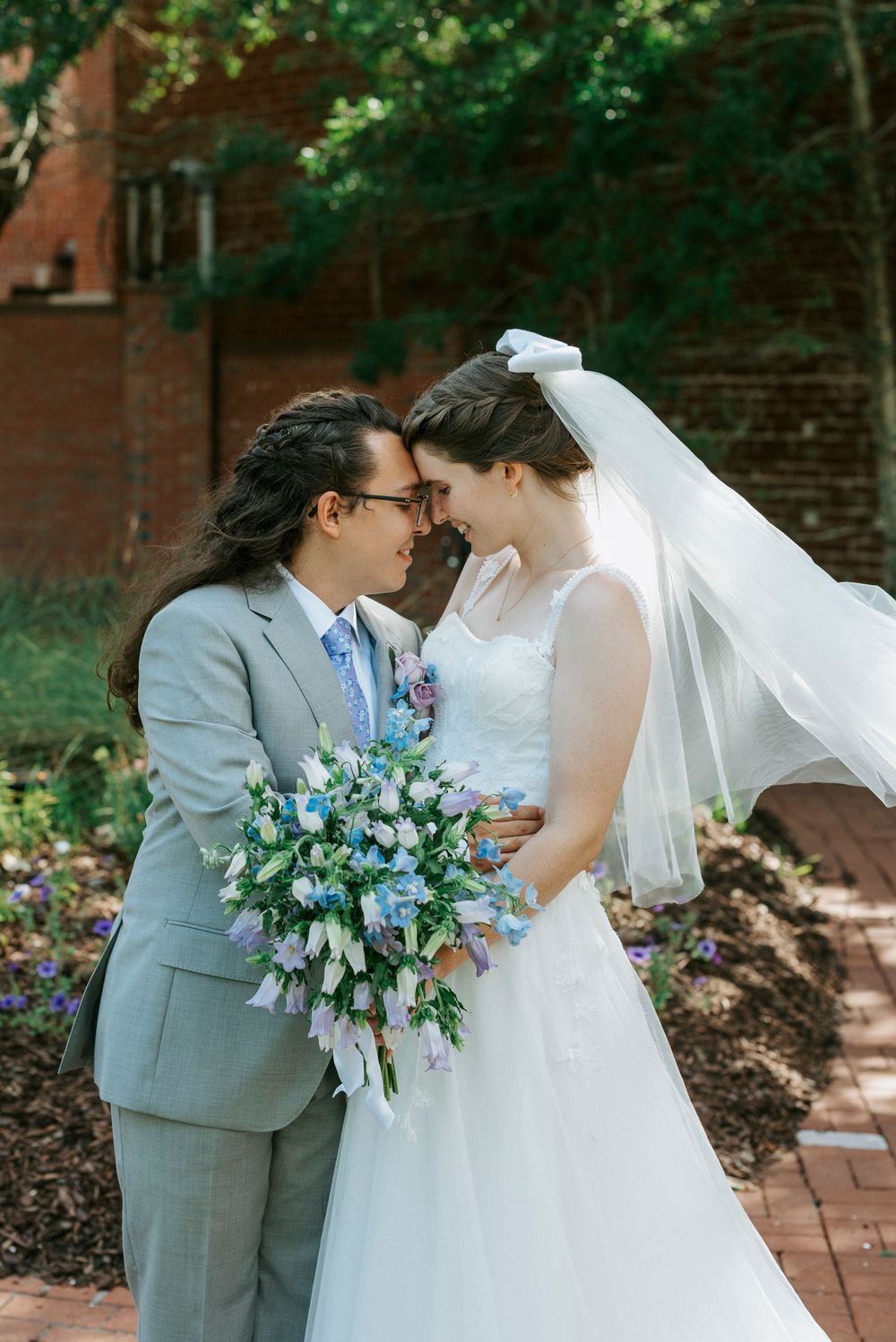 Bride and groom touching foreheads outdoors, bride holding blue and white bouquet, veil flowing in breeze.