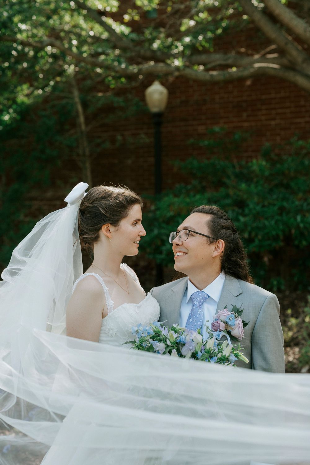 Bride and groom gazing at each other outdoors, veil flowing, holding blue floral bouquet, brick wall background.