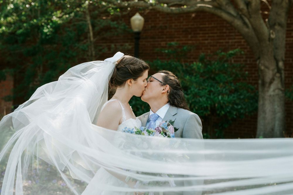 Two brides share a romantic kiss outdoors, with a flowing wedding veil billowing gracefully around them near a brick building.