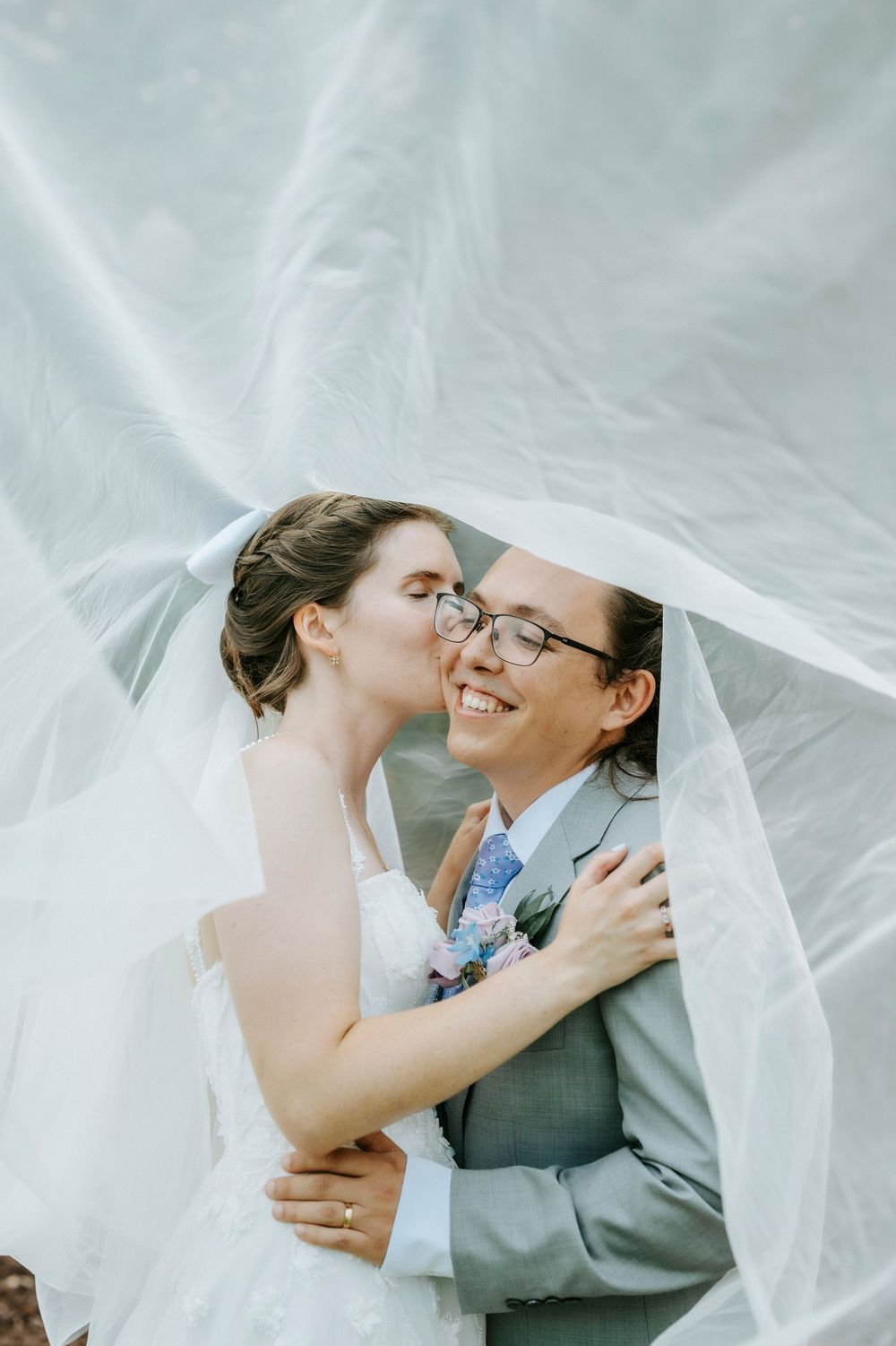 Two brides share a tender kiss under a flowing white veil on their wedding day, one in a suit and one in a gown.