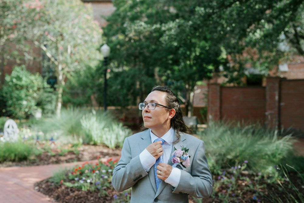 Groom in gray suit adjusting tie in garden setting with lush greenery and brick building backdrop.