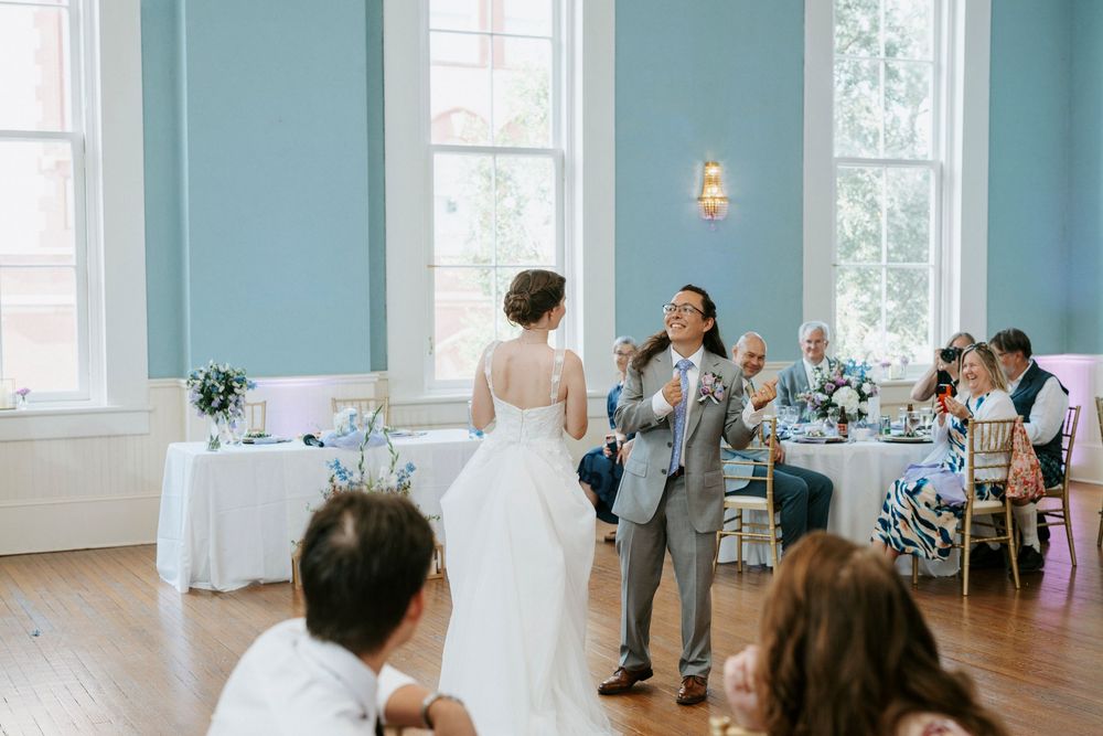 Bride and groom share first dance in elegant blue ballroom with wedding guests seated at decorated tables.