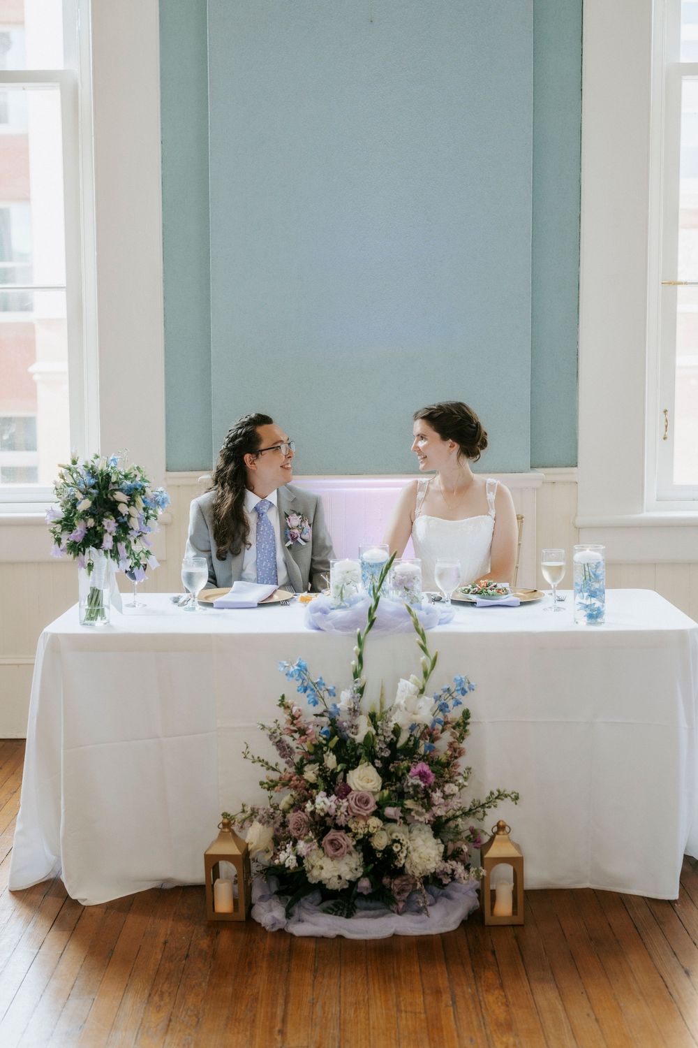 Two brides share a smile at their sweetheart table adorned with blue and white floral arrangements at their wedding reception.