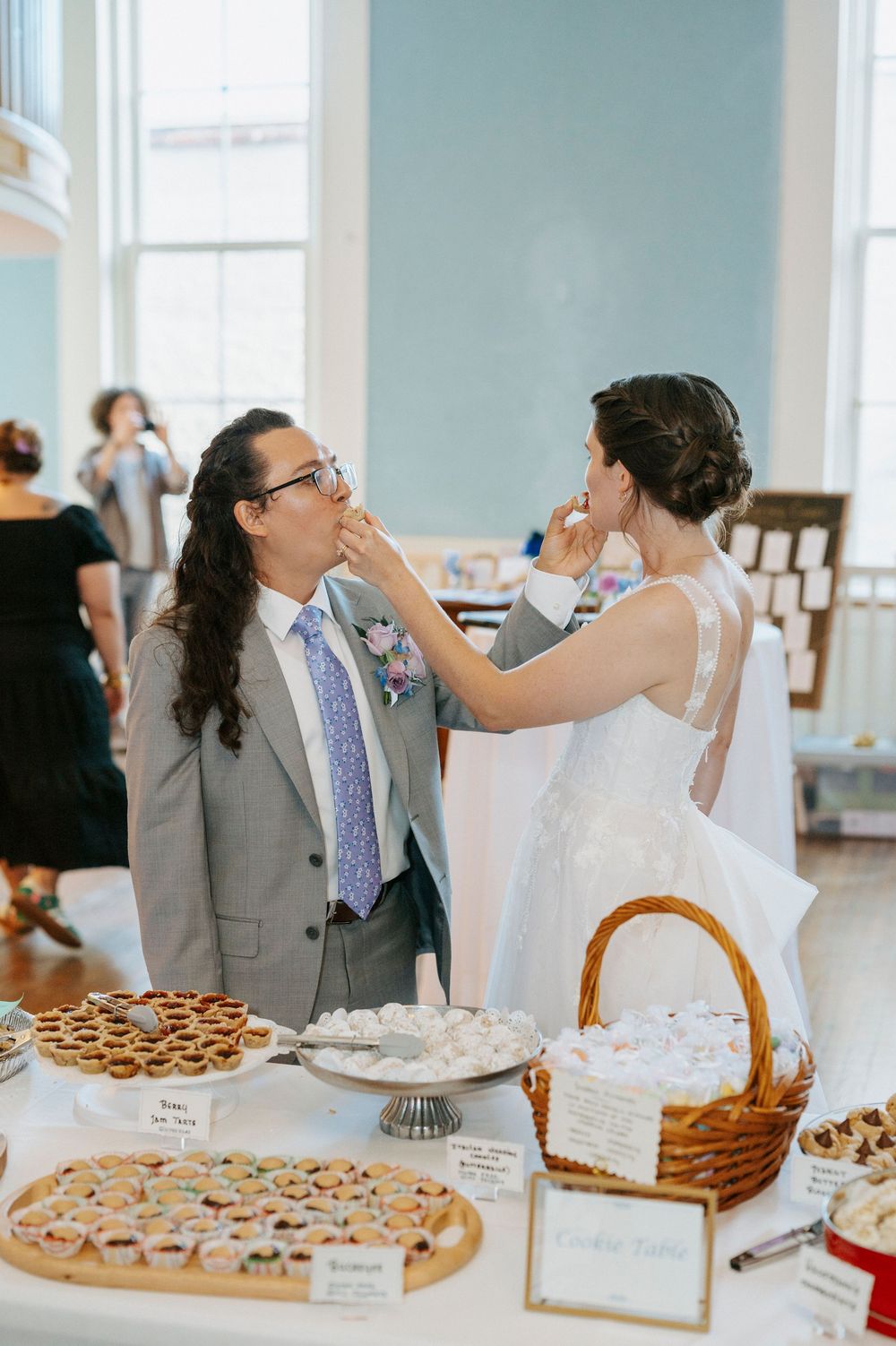 Bride and groom feeding each other cake at a wedding dessert table filled with cookies and pastries.