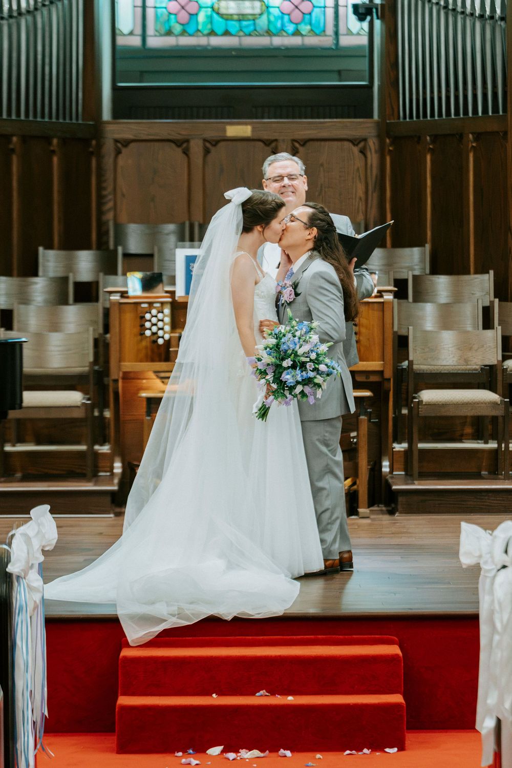 Bride and groom share first kiss at church altar, bride in white gown with veil holding blue bouquet.