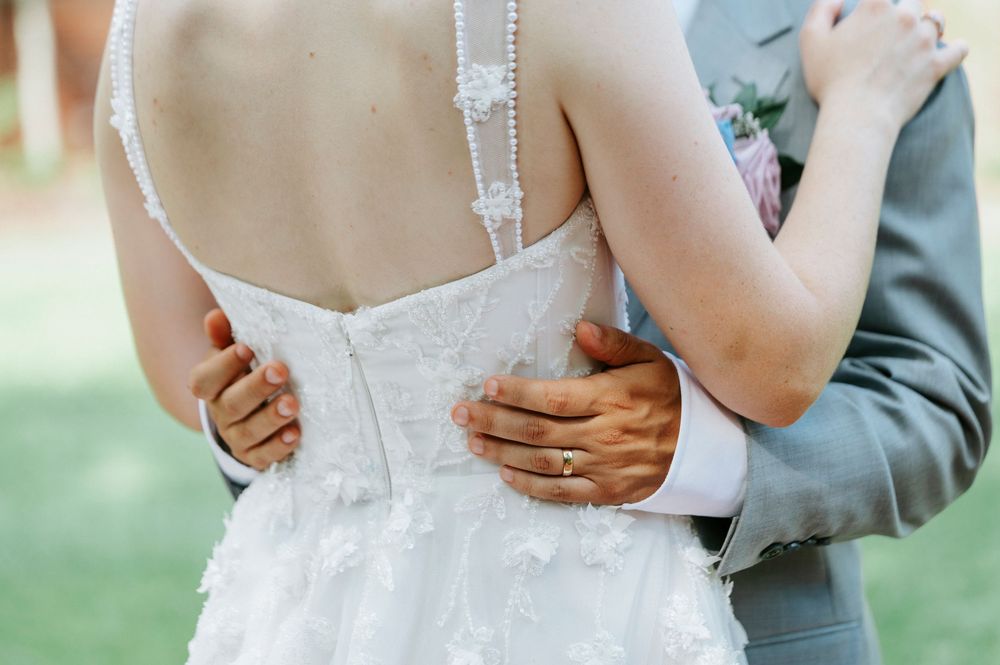 Bride and groom embracing, showing off lace wedding dress back with pearl buttons and diamond engagement ring.