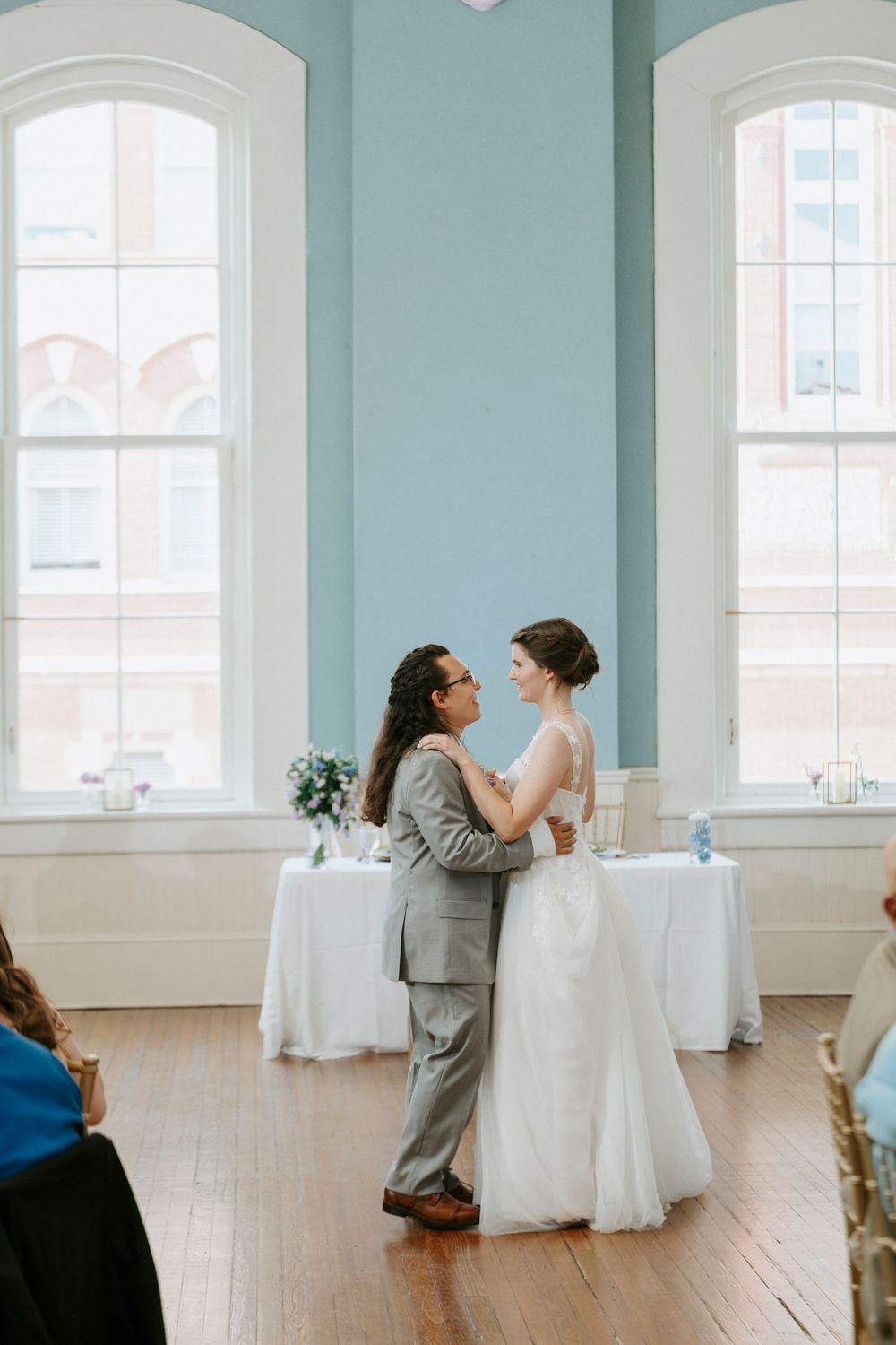 Two brides share their first dance in an elegant blue-walled ballroom with hardwood floors and tall arched windows.