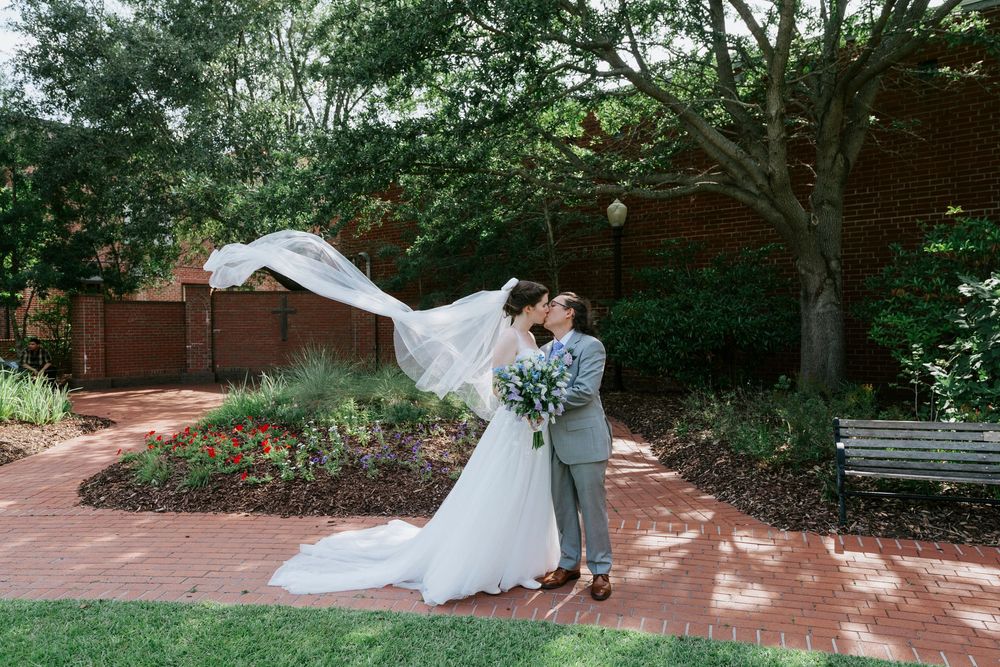 Bride and groom share a kiss outdoors as wedding veil blows in the wind, surrounded by lush garden greenery.