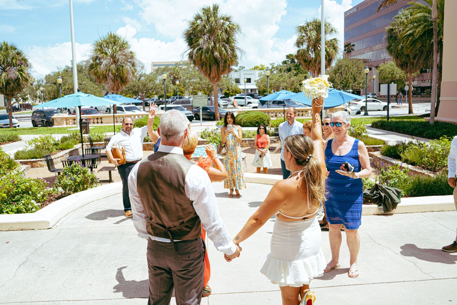 Couple holding hands at outdoor wedding ceremony with guests, palm trees, and sunny Florida backdrop.