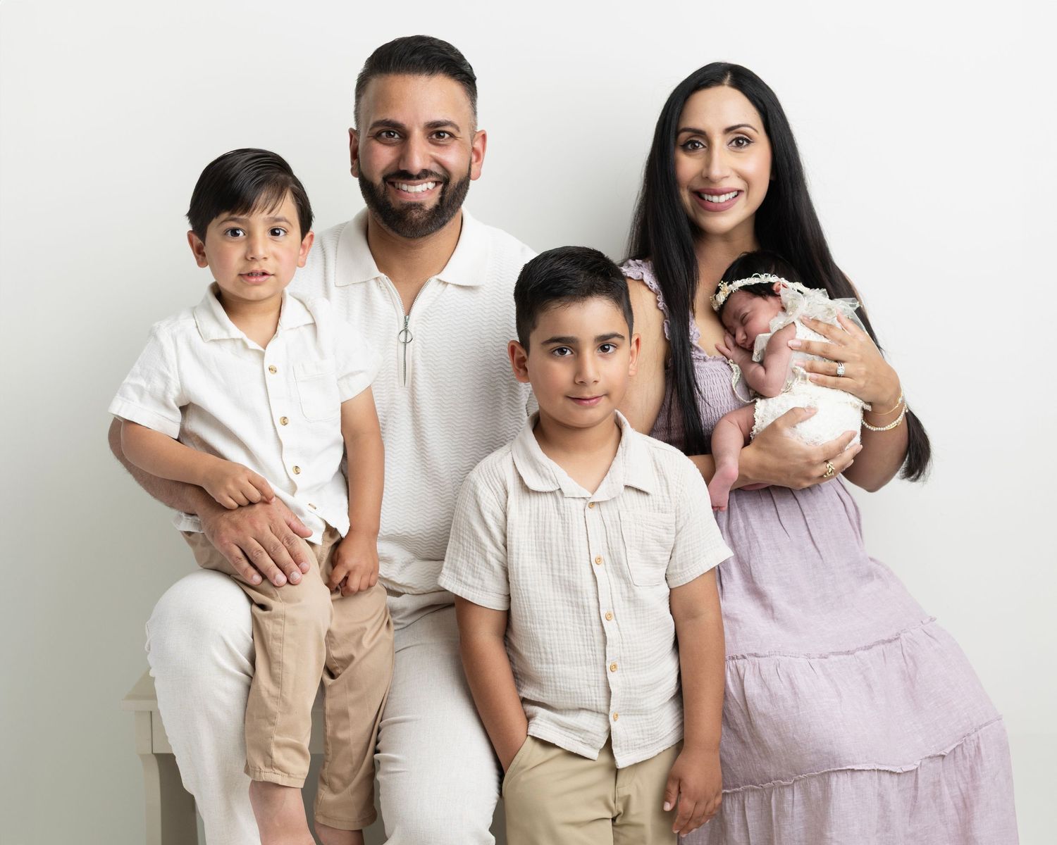 Family portrait with parents, two young boys, and a newborn baby against a white studio background.