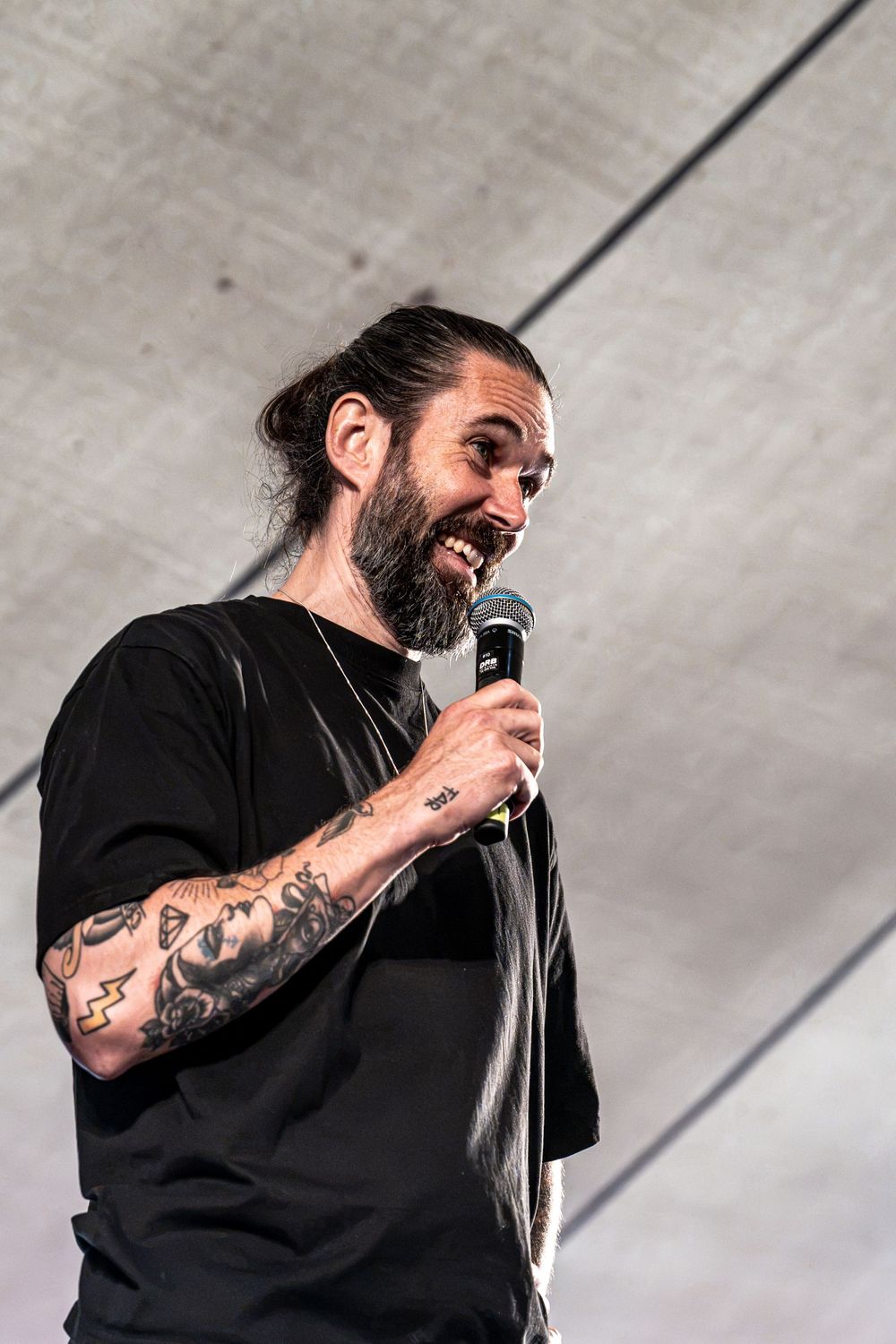 Tattooed man with beard and man bun smiling while holding microphone on stage wearing black t-shirt.
