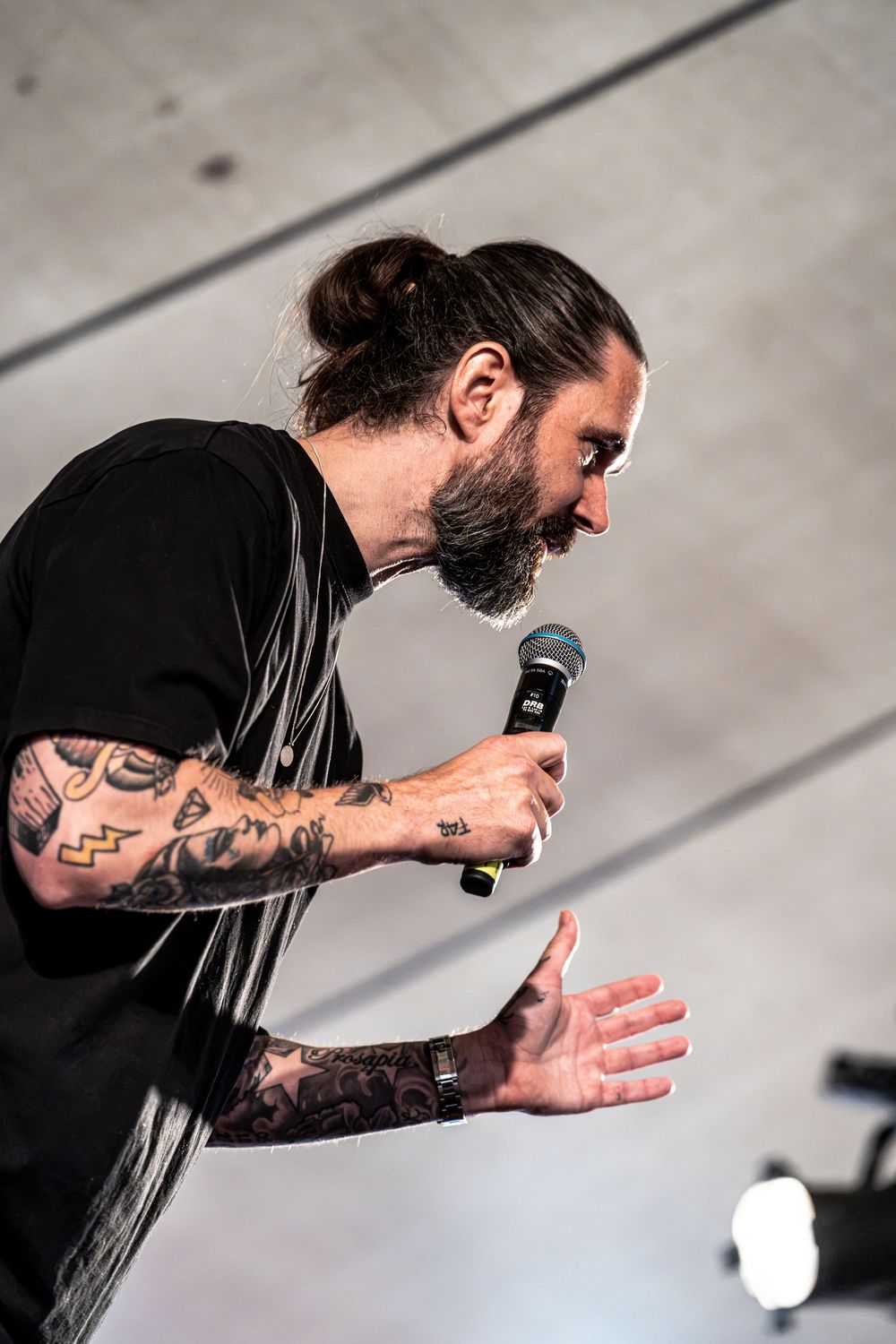 Tattooed male singer with beard and man bun performing on stage, holding microphone in black t-shirt.