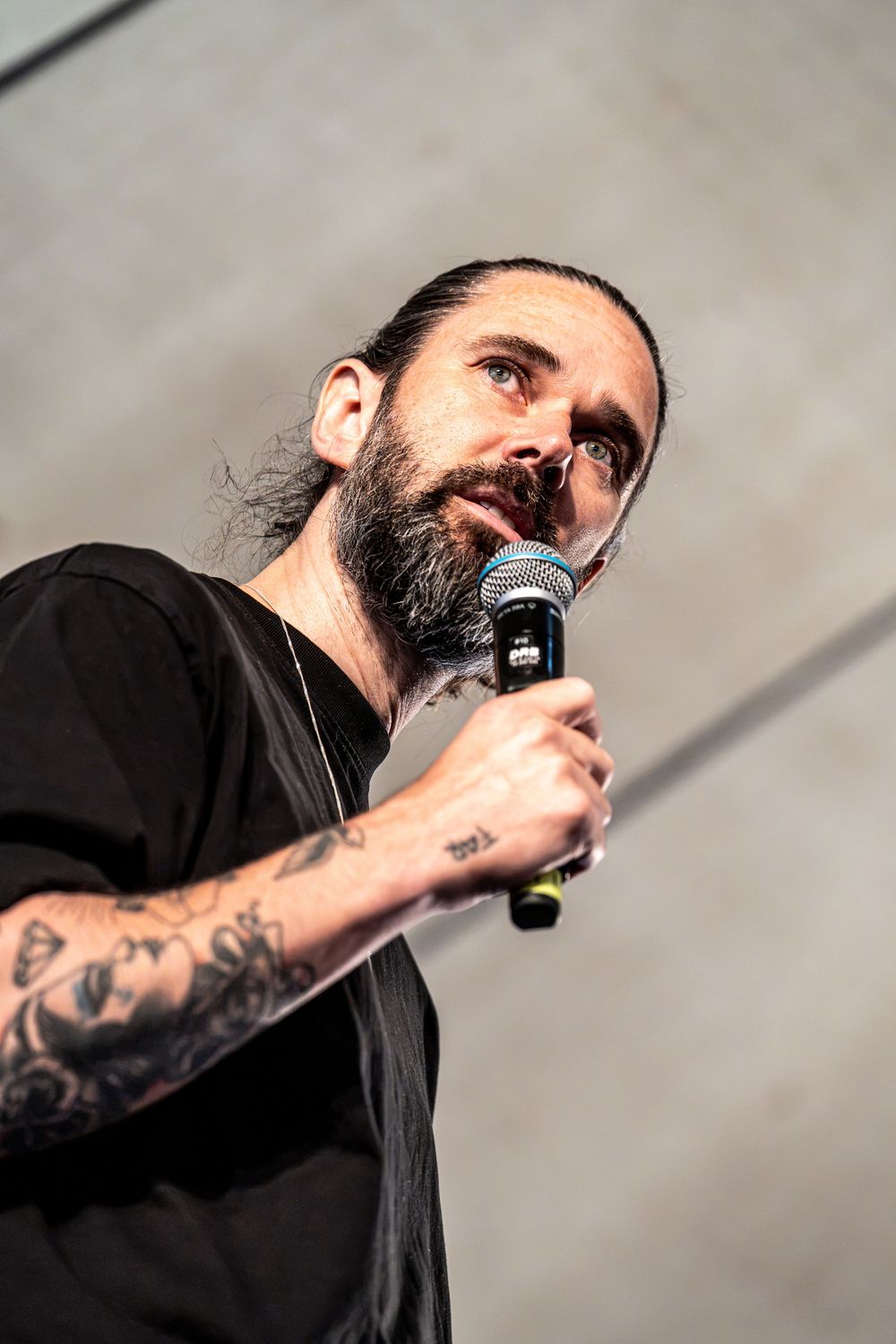 Tattooed man with long hair and beard holding a microphone on stage, wearing a black t-shirt.