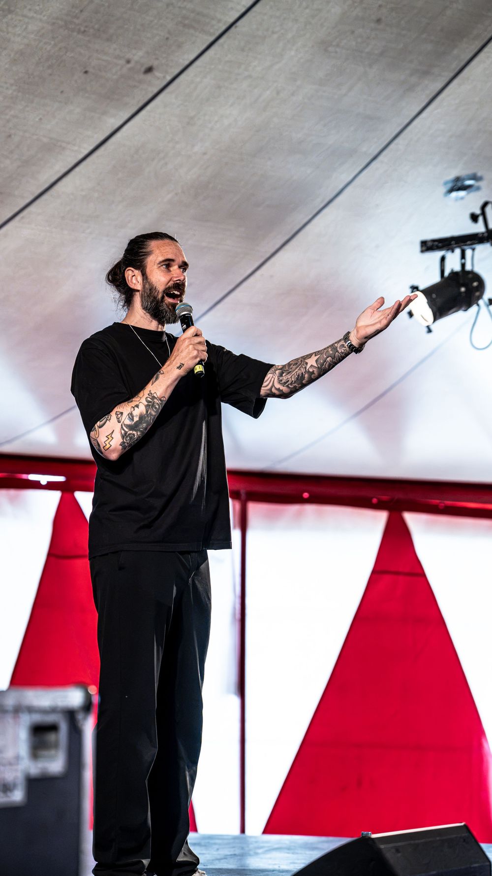 Tattooed man in black outfit speaking into microphone on stage with red triangle backdrop.