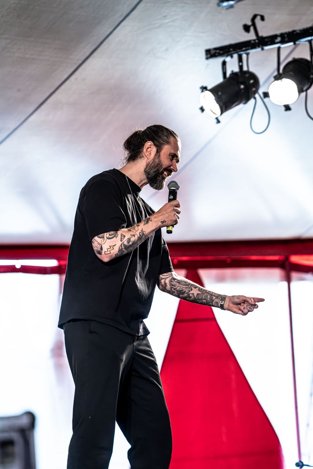 Tattooed male performer in black outfit singing into microphone on stage at outdoor music festival.