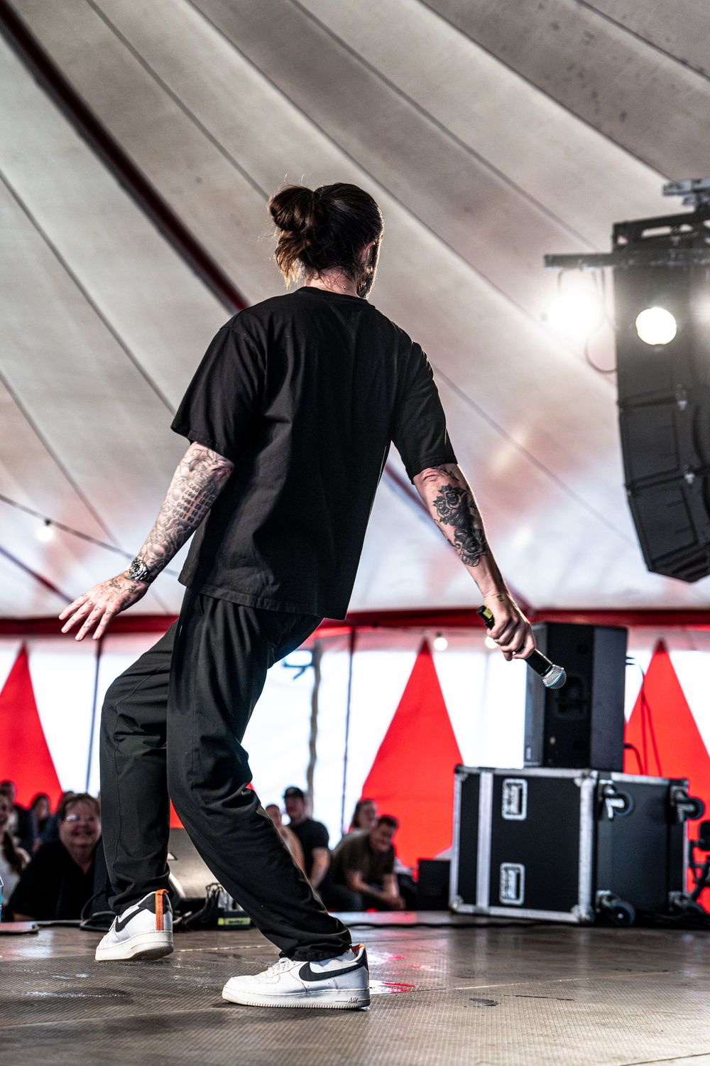 Tattooed performer in black outfit and Nike sneakers holds microphone on stage at outdoor music festival.