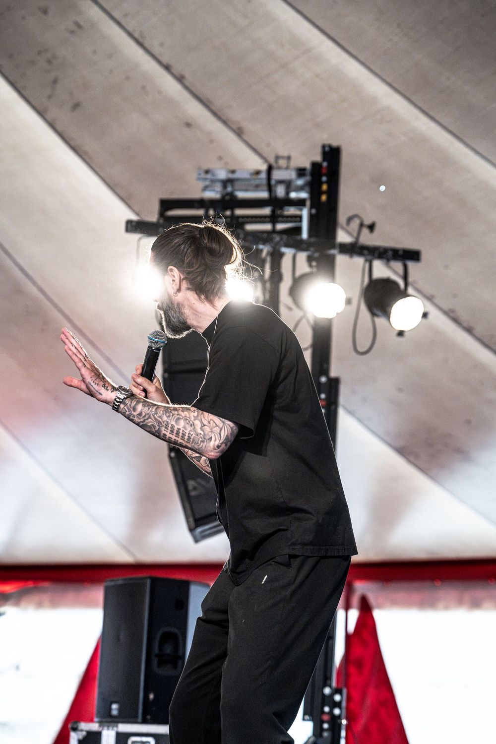 Heavily tattooed male performer in black t-shirt leans forward with microphone under bright stage lights.