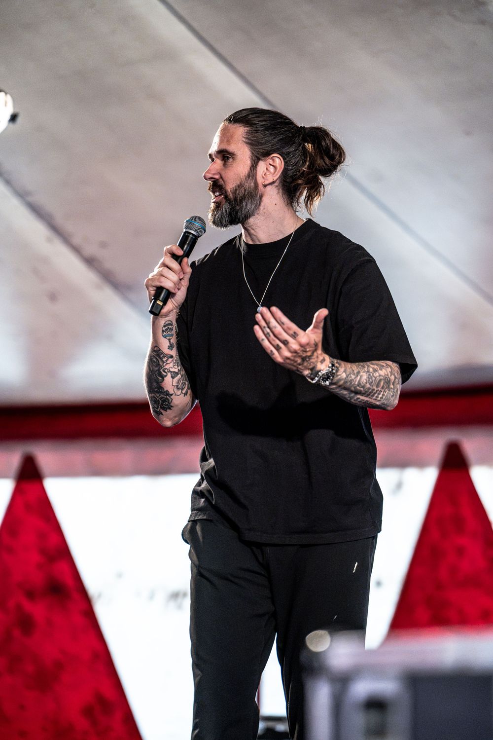 Tattooed male performer in black outfit speaks into microphone on stage with red backdrop.