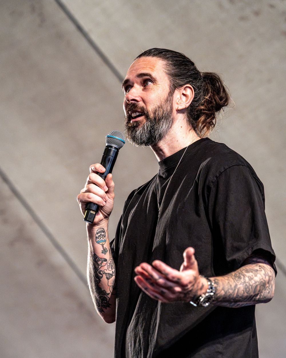 Tattooed man with bun hairstyle and beard speaking into microphone wearing black shirt outdoors.