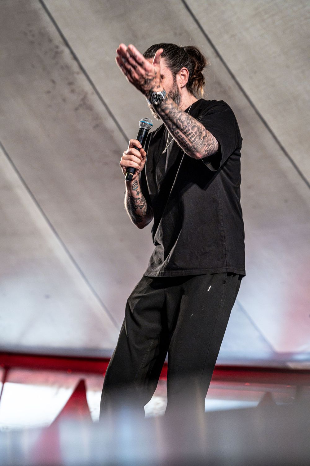 Tattooed male performer in black outfit singing into microphone on stage with hand raised.