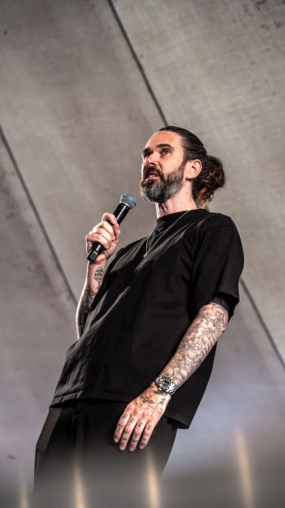 Heavily tattooed bearded male performer singing into microphone on outdoor festival stage.