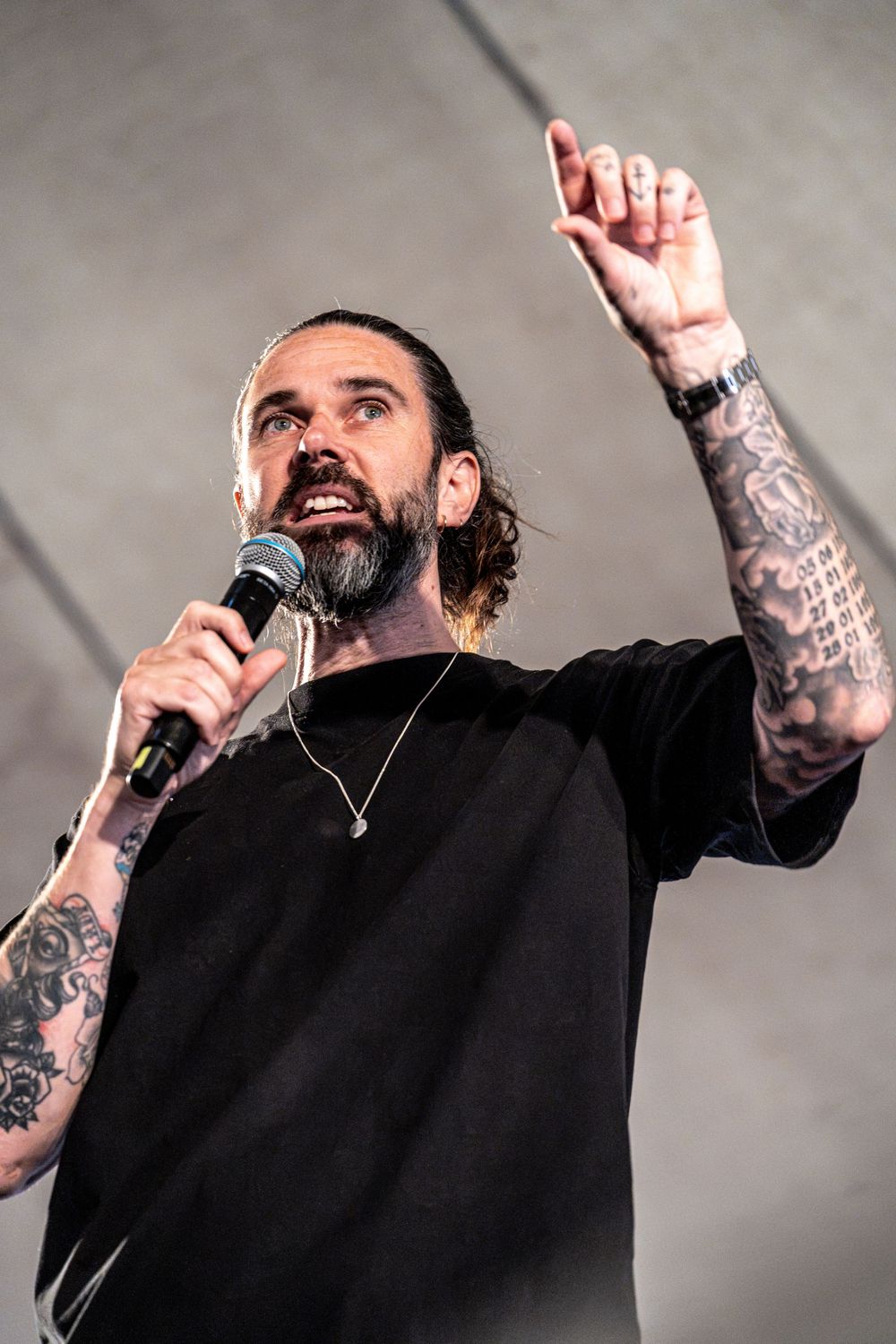 Bearded tattooed male vocalist raises finger while singing into microphone on outdoor stage.