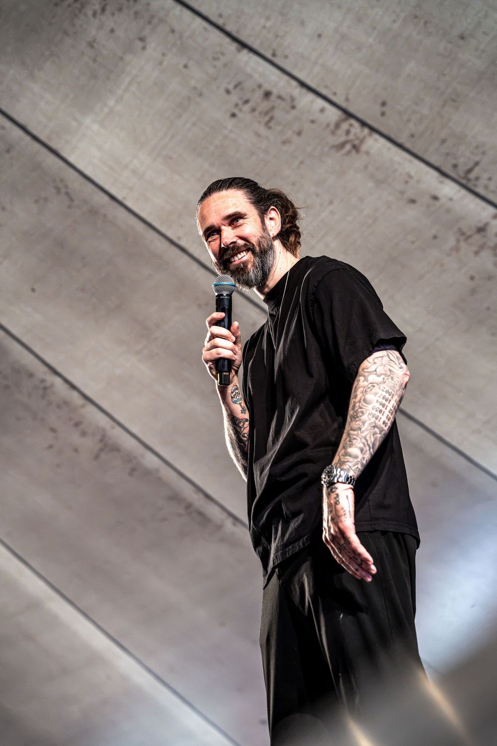 Smiling tattooed man with beard holding microphone on stage, wearing all black outfit, low angle view.