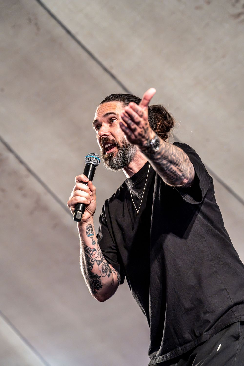 Tattooed man with bun hairstyle holding microphone and pointing finger while performing on stage in black outfit.