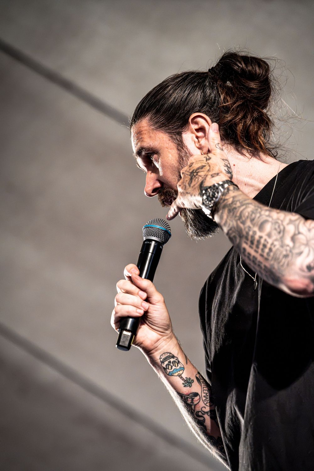Side profile of tattooed bearded performer singing into microphone on stage in black t-shirt.