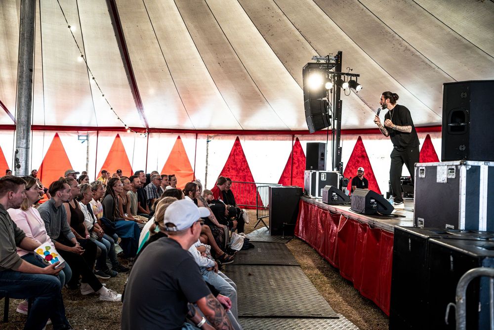 Performer on stage speaking to a seated audience inside a festival tent with red decor.